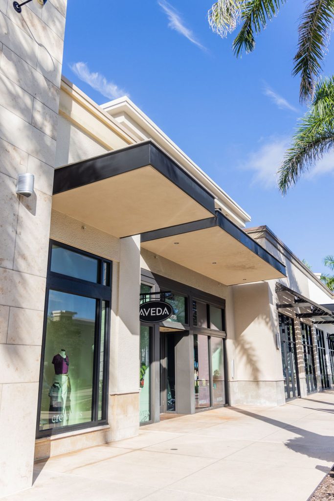 A modern storefront with large windows and a sign reading "Aveda" under a flat awning, this Aveda hair salon—offering hair color, extensions, facials, and nails—is surrounded by palm trees on a sunny day. Discover the best salon Naples FL!. Zen Aveda Salon in Naples, FL
