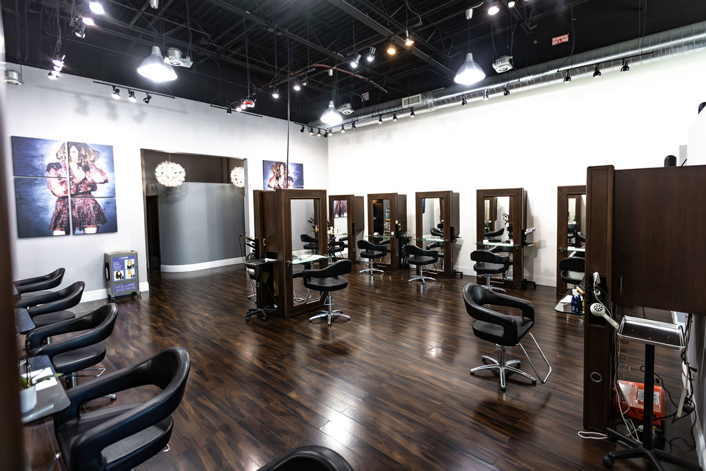 Modern hair salon interior with dark wood floors, black styling chairs, mirrored stations, and artwork on the walls—all beautifully lit. Experience top hair color, extensions, facials, and nails at the best salon Naples FL. Zen Aveda Salon in Naples, FL