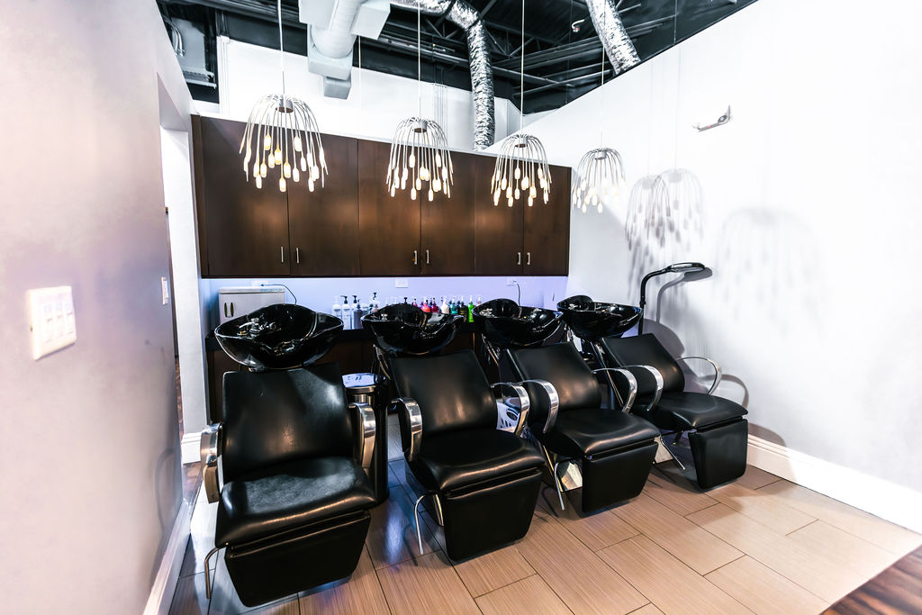 Four black salon shampoo chairs are aligned in front of washing sinks, with overhead lighting and dark cabinetry in a modern, clean interior offering premium spa services Naples, including hair color extensions facials nails. Zen Aveda Salon in Naples, FL