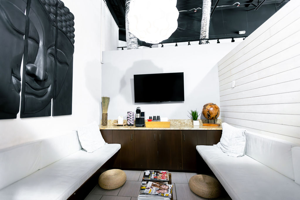 Modern waiting area in this Aveda hair salon features white benches, a TV on the wall, a coffee station, decor accents, and magazines on the floor—perfect for relaxing before enjoying spa services Naples like facials or hair color extensions. Zen Aveda Salon in Naples, FL