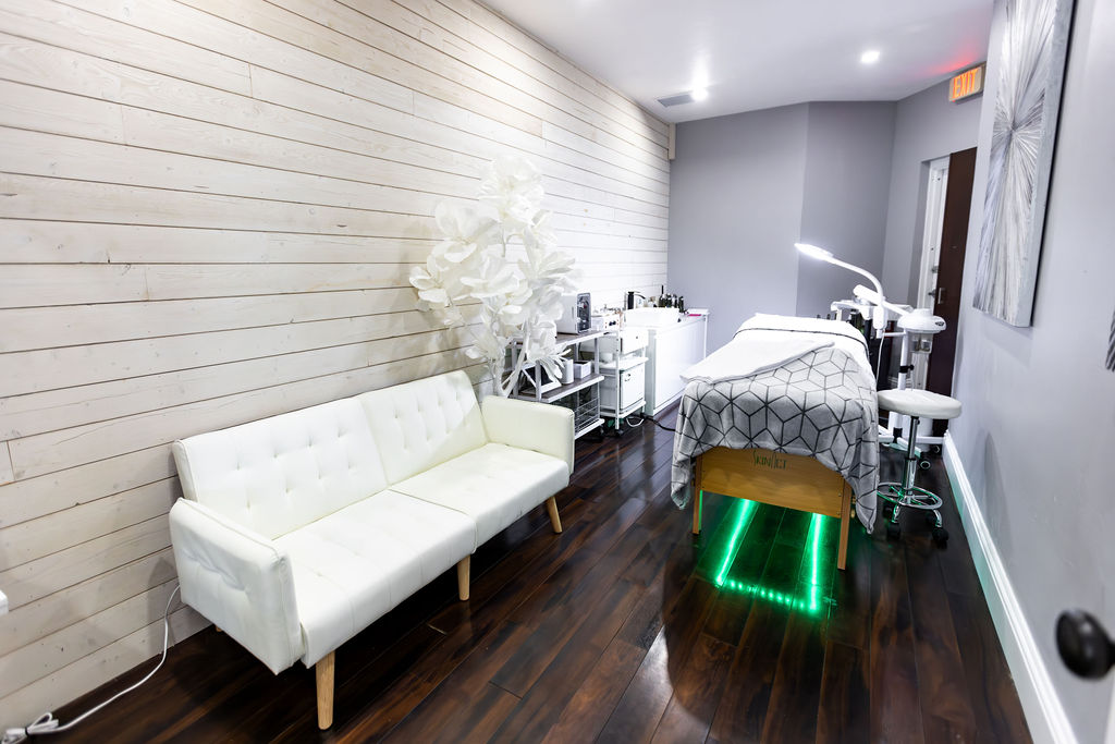 A modern treatment room at this premier salon Naples features a white sofa, wooden walls, dark wood floors, a massage table with a gray cover, and medical equipment along the wall—perfect for facials or Aveda hair salon services. Zen Aveda Salon in Naples, FL