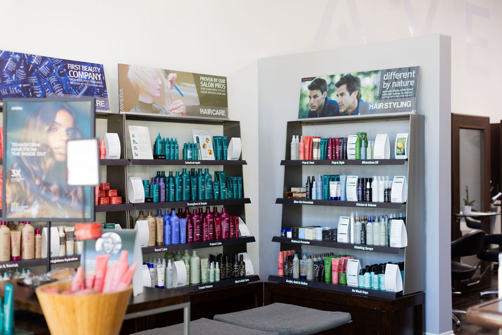 Shelves stocked with various haircare and hairstyling products in a well-lit premier salon Naples, with promotional posters displayed above the products highlighting hair color, extensions, facials, and nails. Zen Aveda Salon in Naples, FL