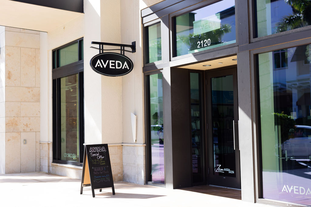 A storefront with an AVEDA sign above the entrance and a sandwich board displaying a special offer outside the door at 2120, highlighting this premier salon Naples as a destination for luxury hair and spa services Naples. Zen Aveda Salon in Naples, FL