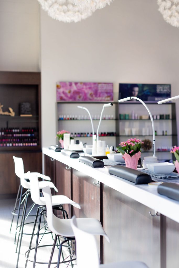 A modern nail salon with white chairs, manicure stations, desk lamps, and pink potted flowers on the counter under natural light. Offering spa services Naples residents love, shelves with nail polish bottles are in the background. Zen Aveda Salon in Naples, FL