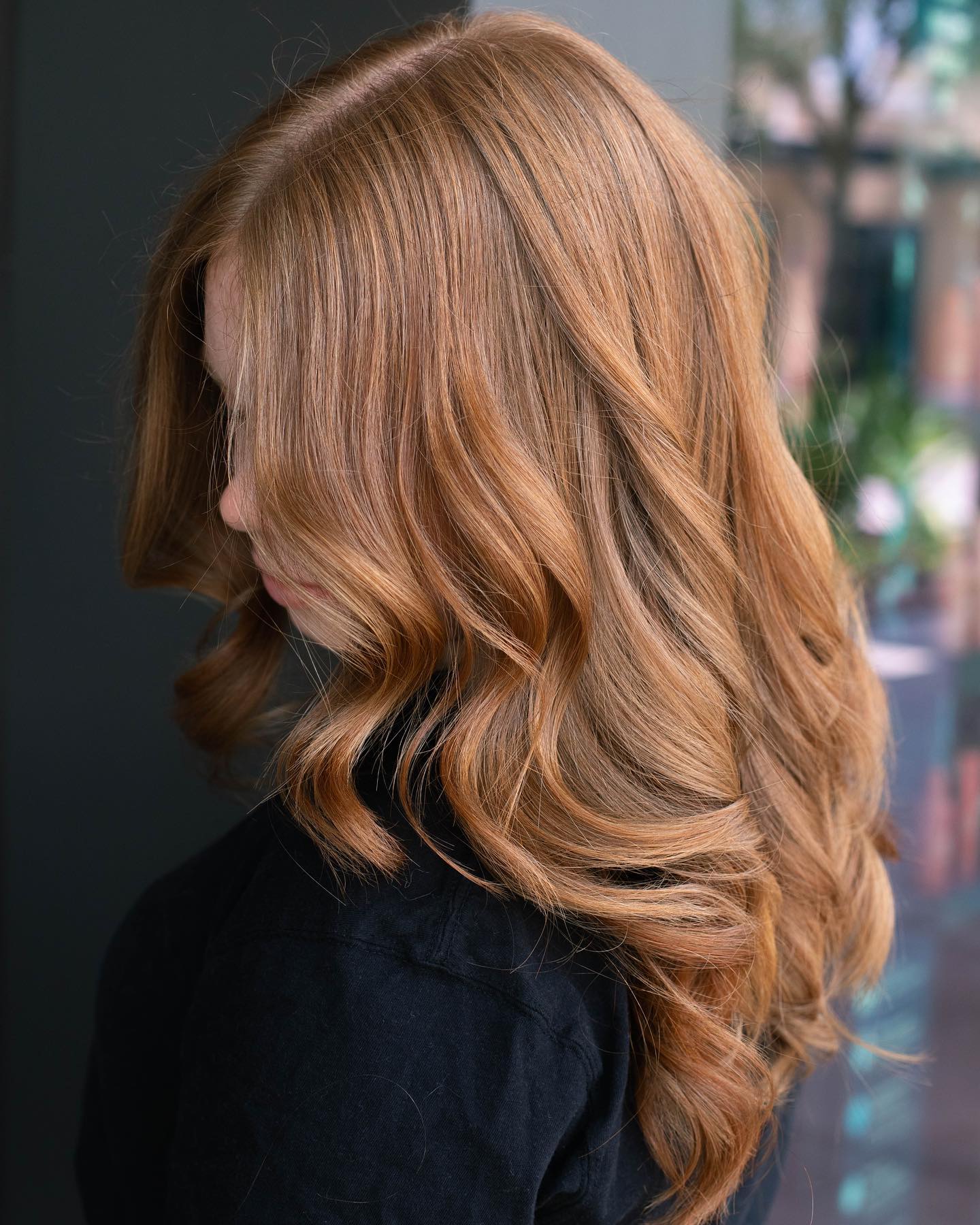 Person with long, wavy, light reddish-blonde hair is shown in profile against a blurred background, wearing a black top—capturing the style you'd find at a premier salon Naples. Zen Aveda Salon in Naples, FL
