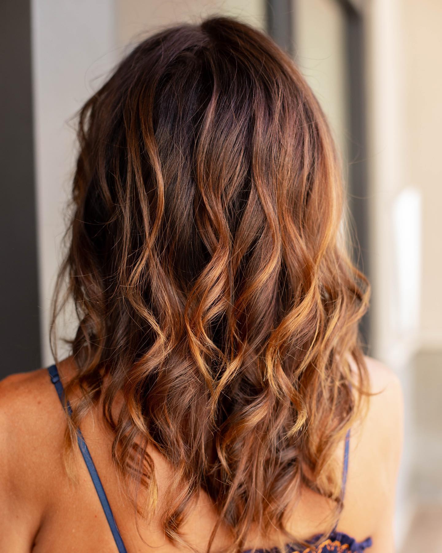 A person with shoulder-length, wavy brown hair with caramel highlights is shown from the back, wearing a spaghetti strap top—showcasing results you’d expect from the best salon Naples FL has to offer. Zen Aveda Salon in Naples, FL
