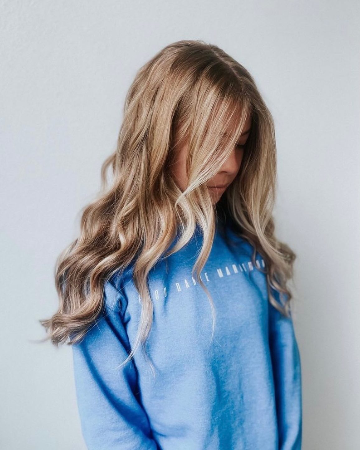 A person with long, wavy blonde hair—styled to perfection as if fresh from the best salon Naples FL—wears a light blue sweatshirt, standing against a plain light-colored wall and looking down. Zen Aveda Salon in Naples, FL