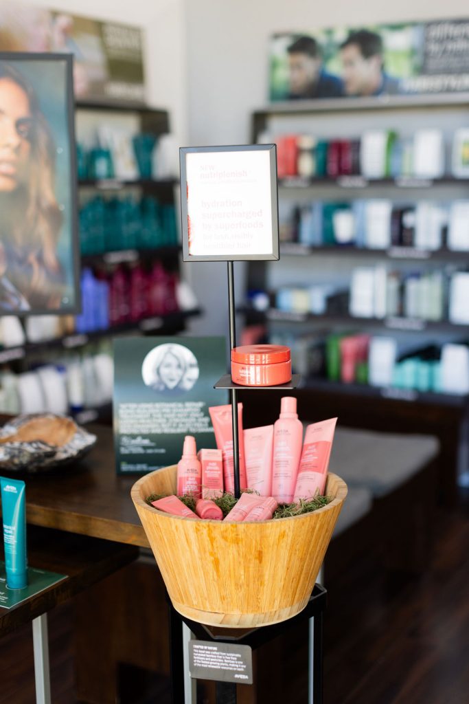 A display stand features a wooden basket filled with pink hair care products, surrounded by shelves of beauty items in a premier salon Naples, offering hair color extensions facials nails and other spa services Naples clients adore. Zen Aveda Salon in Naples, FL