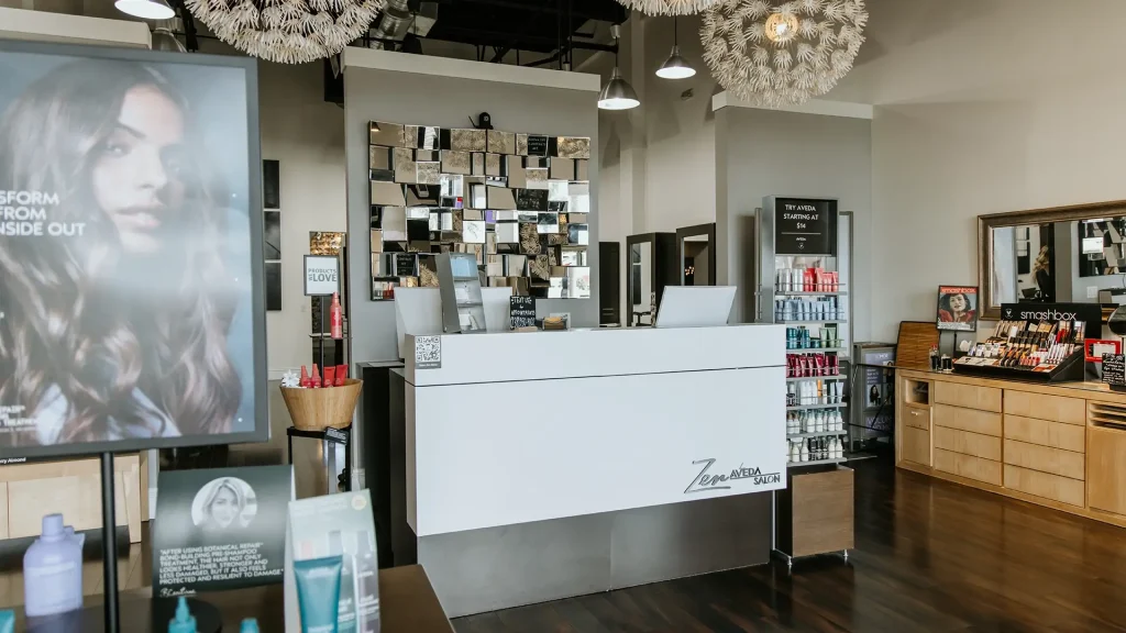 Reception desk at a modern Aveda hair salon with product displays, a decorative mirror, and promotional posters visible in the background—experience the best salon Naples FL has to offer. Zen Aveda Salon in Naples, FL