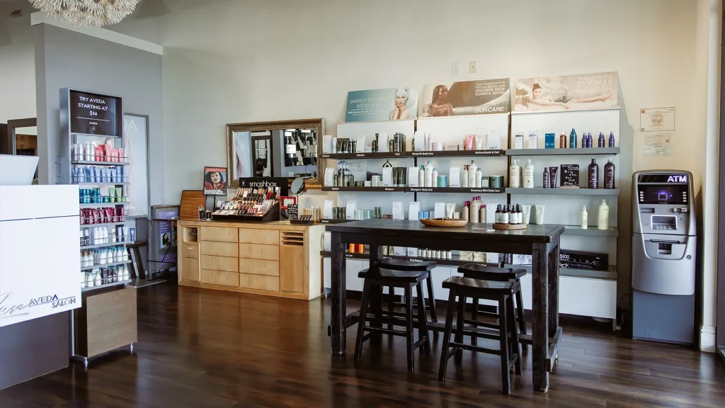 Modern salon retail area with shelves displaying hair and beauty products, a wooden table with stools, a makeup station, and an ATM on the right—experience premier salon Naples services including facials, nails, and Aveda hair color extensions. Zen Aveda Salon in Naples, FL