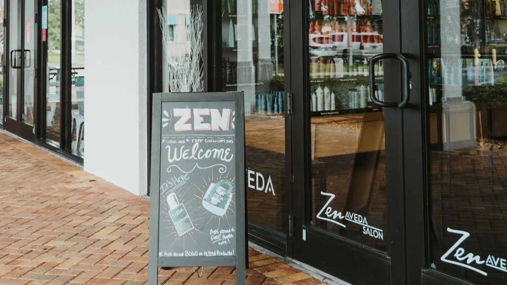 A sidewalk chalkboard sign outside Zen Aveda, a premier salon Naples, displays a welcome message and product drawings next to the salon’s glass entrance doors. Zen Aveda Salon in Naples, FL