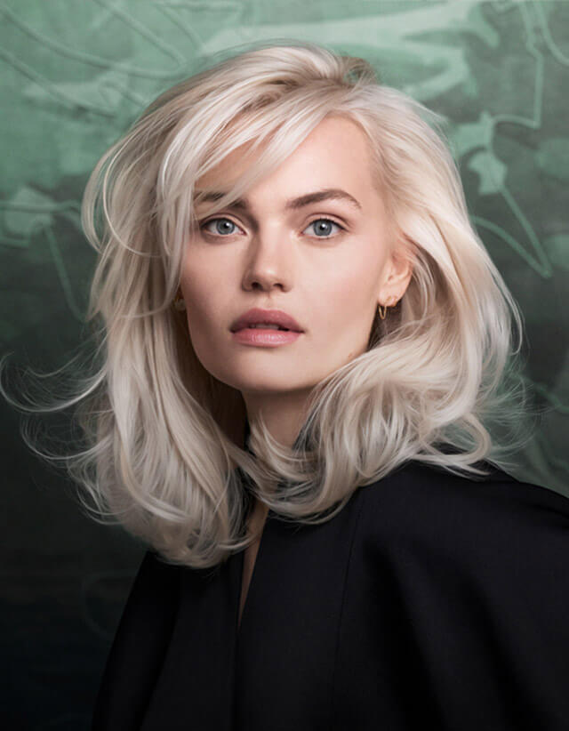 A woman with shoulder-length platinum blonde hair and light eyes poses against a blurred green and black background, showcasing the results of best salon Naples FL hair color extensions facials nails, and wearing a sleek black top. Zen Aveda Salon in Naples, FL