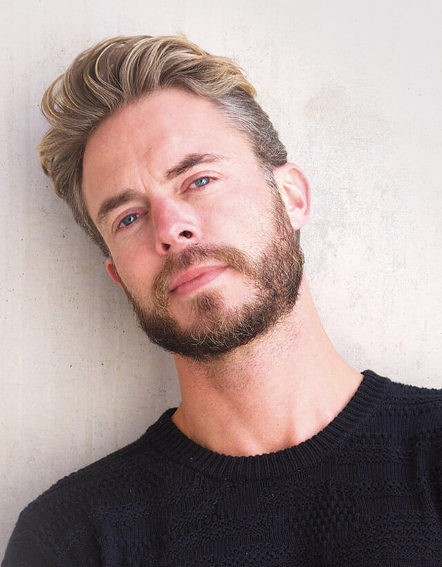 A man with light brown hair and a beard, wearing a black sweater, stands against a light-colored wall, looking slightly to the side—capturing the refined look you’d expect from the best salon Naples FL has to offer. Zen Aveda Salon in Naples, FL